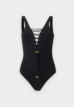 Γυναίκα ακάλυπτη, φοράει μαύρο ολόσωμο μαγιό με δέσιμο μπροστά, σε απλό λευκό φόντο.