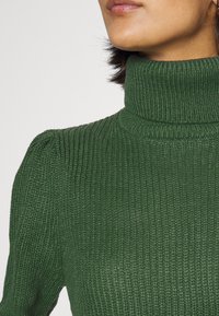 Pull à col roulé vert côtelé avec un tissu tricoté texturé et des manches légèrement bouffantes discrètes. Gros plan sur les détails du col montrant un col montant ajusté.