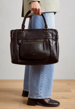 Person trägt hellblaue Jeanshose und schwarze Absatzschuhe und hält eine dunkelbraune Ledertasche mit Reißverschlusstasche vorne.