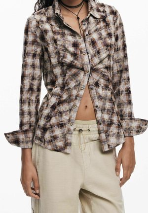 Femme portant une chemise à carreaux marron et beige déboutonnée sur un pantalon beige avec lien coulissant, avec des chokers noirs superposés autour du cou.