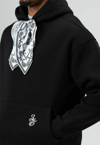 Zwarte hoodie met een kangoeroezak, voorzien van een witte bandana-accent geknoopt bij de hals en een klein wit geborduurd logo aan de voorkant.