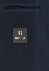 Dunkelblaues Jackenfutter mit einem schwarzen Stoffetikett, auf dem "B BOGGI MILANO" steht, nahe einem Schlitz der Innentasche eingenäht.