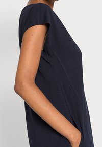 Sininen lyhythihainen paita, joka on valmistettu kevyestä materiaalista ja jossa on väljä istuvuus, sivutaskut ja hienovaraiset ompeluyksityiskohdat sivuilla.