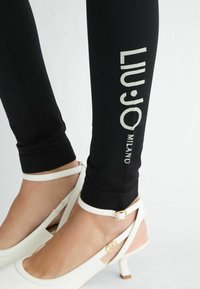 Leggings negros con el logo "LIU·JO MILANO" en plata en el lateral, combinados con zapatos de tacón blancos con tiras que presentan un detalle dorado.