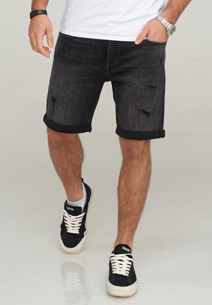 Black denim shorts with a rolled hem, featuring a distressed texture and a slim fit. Worn with black sneakers, showcasing a casual style.