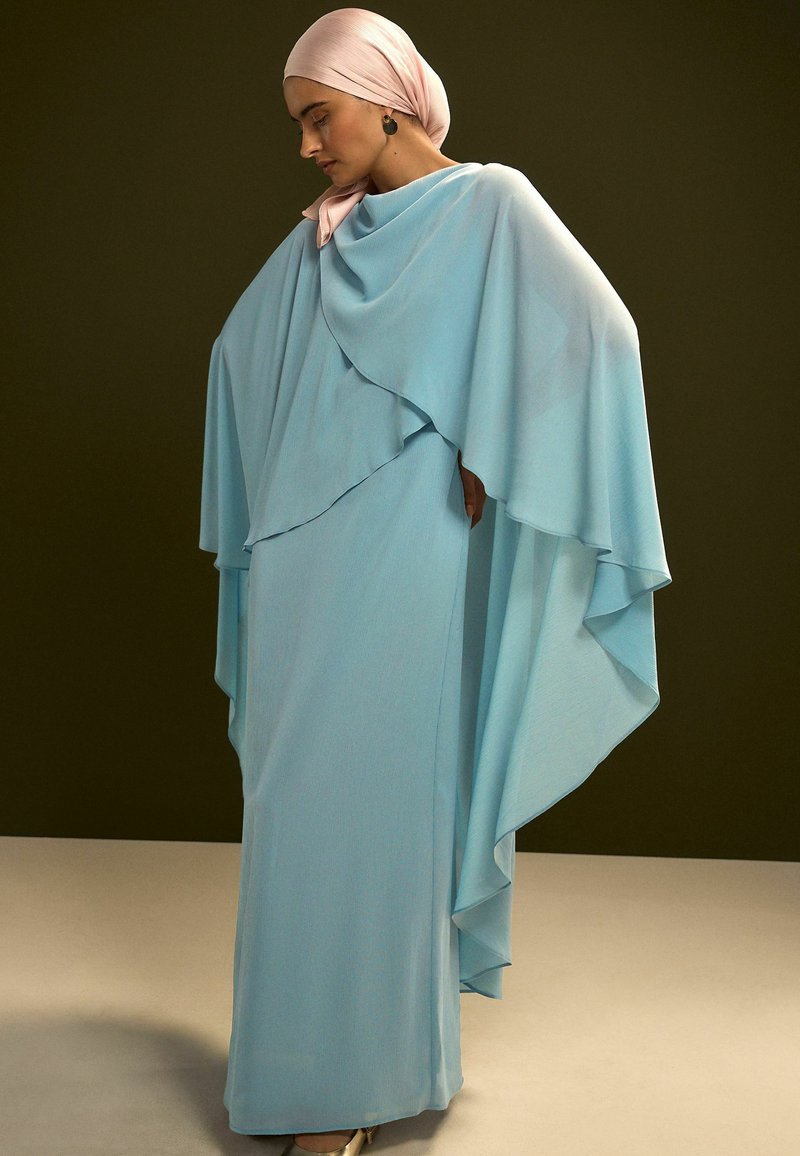 Vestido con capa fluida de color azul claro, diseño en capas y textura suave. Acompañado de un pañuelo rosa en la cabeza y pendientes sutiles.