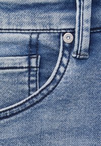 Gros plan sur un jean en denim bleu clair, avec une poche visible, des coutures contrastées, un détail de bouton métallique et une texture douce.