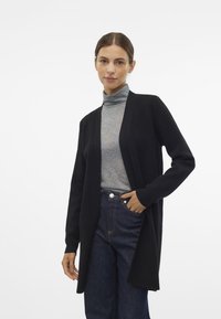 Cardigan nero con design aperto, realizzato in tessuto morbido. Indossato sopra un dolcevita grigio, abbinato a jeans in denim scuro a vita alta.