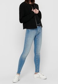 Veste noire courte avec un col arrondi, associée à un jean skinny bleu clair et des baskets blanches, debout devant un fond neutre.