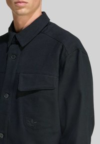 Detailný záber na osobu oblečenú v čiernej košeli na gombíky s vreckom na klopu a nenápadným logom Adidas na hrudi.