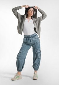 Szara bluza rozpinana na zamek, biały dopasowany top, niebieskie spodnie typu cargo z elastycznymi mankietami oraz lekkie trampki z zielonymi akcentami. Wyrazisty, codzienny zestaw.