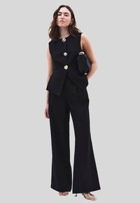 Zwarte mouwloze top met gouden knopen, gecombineerd met vloeiende zwarte broek. Het model houdt een zwarte clutch vast en draagt metalen haksandalen.