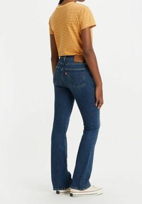Los jeans de mezclilla azul acampanados presentan una cintura de altura media y bolsillos traseros. El modelo lleva una camisa de manga corta a rayas amarillas y blancas.