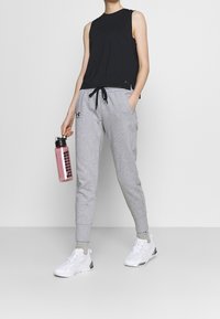 Person som bär en svart ärmlös topp, grå sweatpants, vita sneakers, håller en rosa vattenflaska i höger hand, står mot en enkel bakgrund.