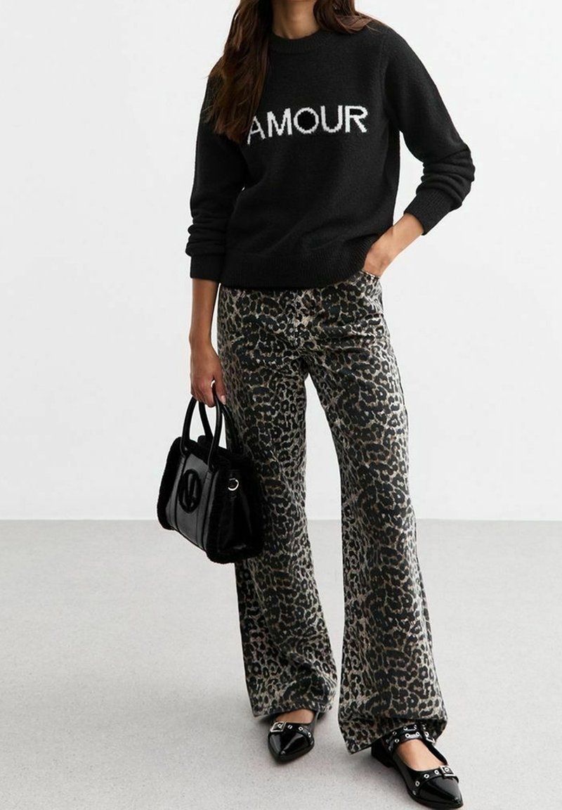 Femme portant un pull noir avec le texte "AMOUR", pantalon à imprimé léopard, sac à main noir et chaussures noires pointues, debout sur un sol clair.