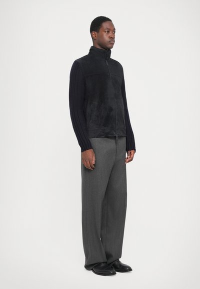 Gilet zippé noir avec des manches texturées contrastantes, associé à un pantalon gris sur mesure. Tissu lisse sur le corps, détails en tricot sur les manches.