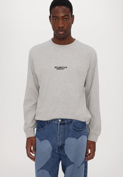 Gray long-sleeve shirt with "SELF LOVE CLUB OPEN 24/7" text in black; paired with blue jeans featuring large heart-shaped patches.