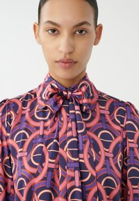 Paarse zijden blouse met cirkelvormige roze en oranje patronen, een strikdetail bij de hals en geplooide schoudernaad. Soepele textuur en levendige kleuren.