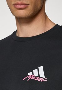 Sort T-shirt lavet af bomuld, med et hvidt Adidas-logo og pink "Messi" tekst. Rundhalsen er standard, med en glat tekstur.