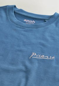 Blå crewneck-trøje med "Picasso" broderet i pink på brystet og "apoh London" trykt indeni kraven.