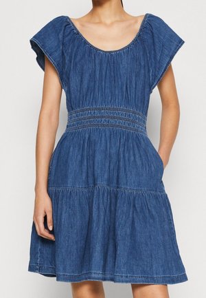 Woman wearing short-sleeve blue denim dress with gathered waist and tiered skirt, hands resting by sides, plain background.