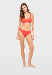 Femme debout pieds nus, portant un soutien-gorge rouge corail et un sous-vêtement style bikini assorti sur un fond uni clair.