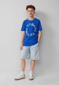 T-shirt en coton bleu avec le texte graphique blanc "L'AVENIR D'AUJOURD'HUI", associé à des shorts en denim clair et des baskets blanches avec des semelles beiges.