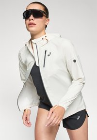 Letvægts hvid løbejakke med justerbar hætte, sorte detaljer og lynlås-lomme. Sammen med en sort top og shorts.