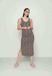 Mulher vestindo um vestido midi preto floral sem mangas, com uma fenda lateral, óculos verdes e sandálias de plataforma bege, em pé contra um fundo liso.