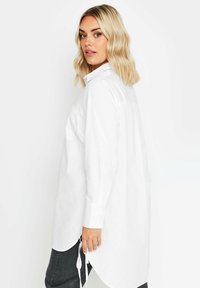Camisa blanca de manga larga con cuello, dobladillo redondeado y lazos laterales ajustables. Fabricada en una tela ligera, presenta un corte relajado.