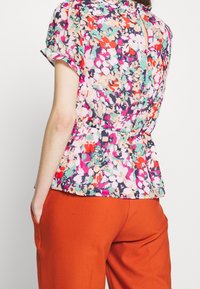 Blusa floral con mangas cortas enrolladas, diseño peplum, patrón multicolor de rosa, verde y púrpura, con un detalle de ojal en la espalda.