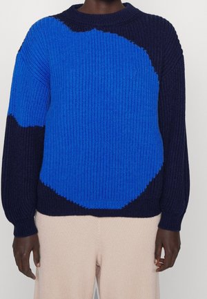 Personne portant un pull en tricot épais avec un grand motif abstrait bleu sur un fond marine et un pantalon beige à rayures côtelées.
