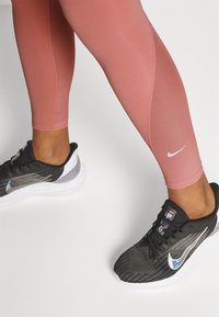 Leggings deportivos rosas con una textura suave, combinados con zapatillas negras y blancas que tienen una parte superior texturizada y el swoosh de Nike reflectante.