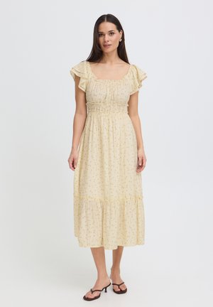 Femme debout portant une robe midi jaune clair avec des manches à volants et un petit imprimé floral, associée à des sandales à talons noires.