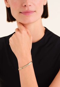 Gouden metalen armband met een geweven ontwerp, gedragen om een pols, met een gladde, glanzende afwerking en een slanke profield.
