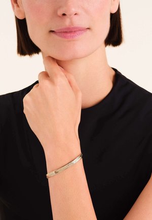 Gouden metalen armband met een geweven ontwerp, gedragen om een pols, met een gladde, glanzende afwerking en een slanke profield.