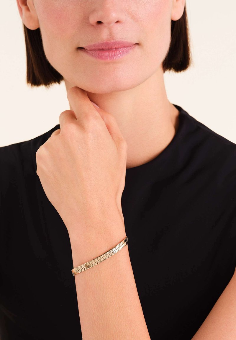 Gouden metalen armband met een geweven ontwerp, gedragen om een pols, met een gladde, glanzende afwerking en een slanke profield.