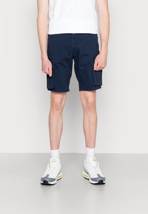 Osoba stojąca w pomieszczeniu, ubrana w granatowe szorty cargo, biały t-shirt, białe skarpetki oraz białe sneakersy z żółtymi i szarymi akcentami.