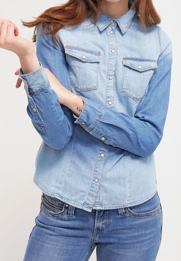 Femme portant une chemise en denim bicolore avec des poches poitrine boutonnées et un jean bleu, les bras croisés à la taille montrant un petit tatouage au poignet.
