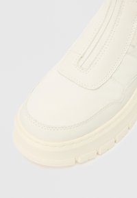 Nærbilde av fronten og siden av en kremfarget sko med tykk såle, synlige sømmer og en glidelås på den øvre panelen.