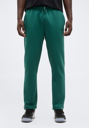 Άνδρας που φοράει πράσινο φόρμα με κορδόνι και μαύρα αθλητικά παπούτσια, στέκεται μπροστά σε ένα απλό φωτεινό φόντο.