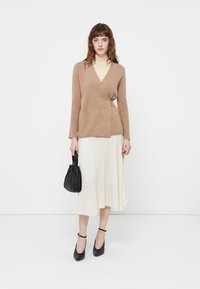Brown wrap sweater in soft knit, paired with a pleated cream midi skirt. Black handbag and black ankle-strap heels complete the look.