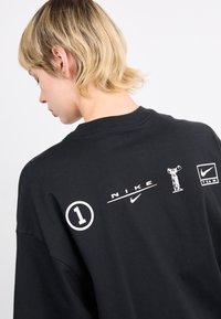 Juodas didelio dydžio t-shirt'as su baltais spaudiniais, įskaitant tekstus ir logotipus, nugaroje. Pagamintas iš medvilnės, su glotnia tekstūra.