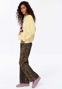 Žena s kudrnatými vlasy, oblečená v žlutém květinovém svetru, leopardích kalhotách a růžových teniskách, stojící bokem s jednou rukou v kapse.
