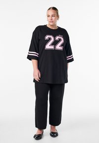 Black oversized t-shirt with pink number "22" and "MILAN" text; features short sleeves with pink stripes. Paired with black pants and flats.