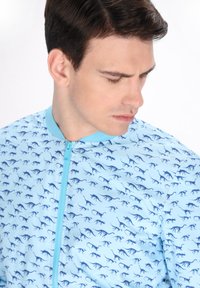 Giacca a zip di colore blu chiaro con un motivo di silhouette di dinosauri blu scuro. La giacca presenta un colletto blu a tinta unita e una texture morbida.