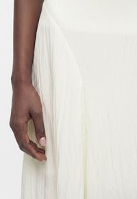 Main à la peau foncée avec des ongles manucurés reposant à côté d'une jupe ou robe en tissu blanc froissé sur un fond clair.