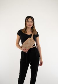 Borsa a tracolla beige con superficie texturizzata e tracolla regolabile, tenuta da una persona che indossa un top nero e pantaloni. Design semplice, senza pattern.