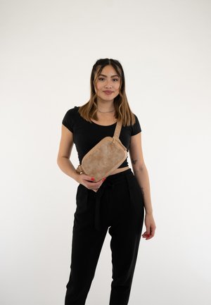 Borsa a tracolla beige con superficie texturizzata e tracolla regolabile, tenuta da una persona che indossa un top nero e pantaloni. Design semplice, senza pattern.