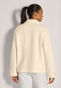 Vrouw met een donkere paardenstaart, gekleed in een crèmekleurige wollen jas en witte broek, met de rug naar de camera gekeerd tegen een effen lichtgrijze achtergrond.